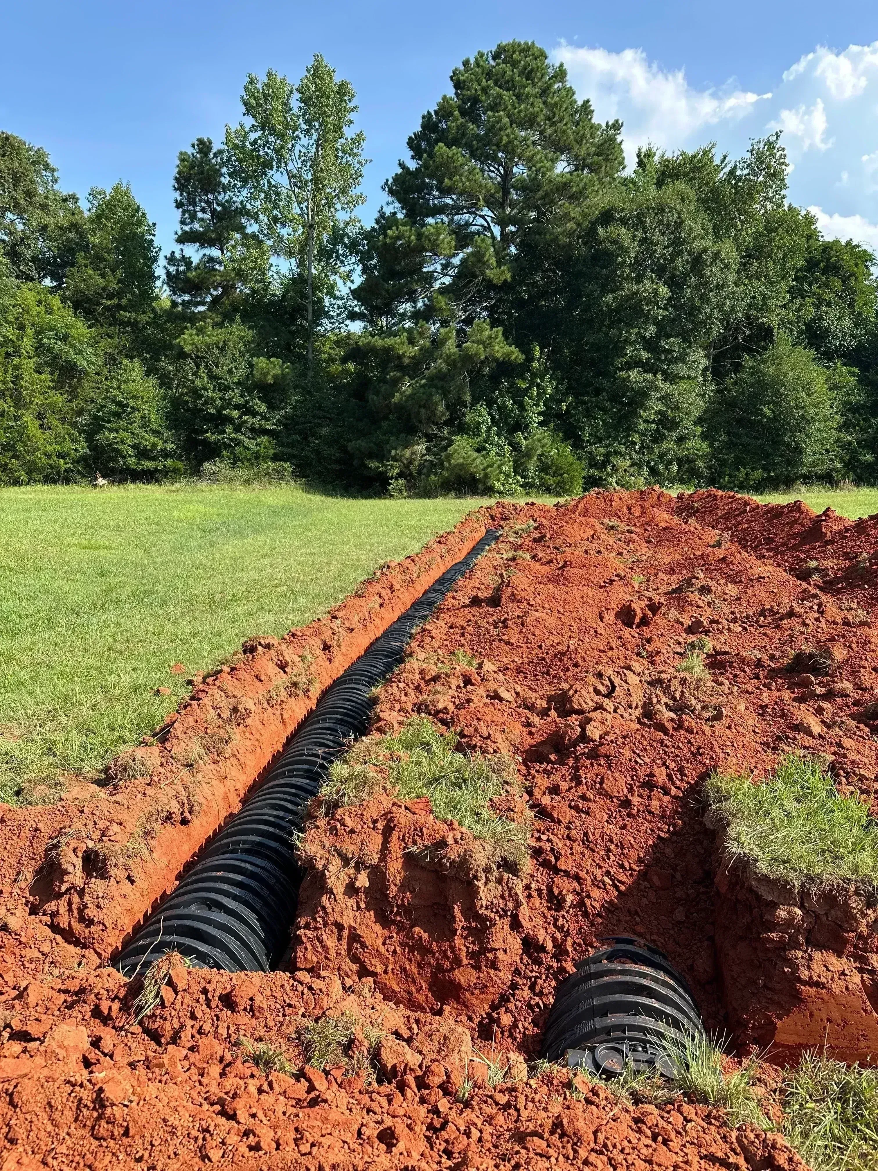 Corrugated drainage pipe installed in excavated trench with red clay soil and grass borders