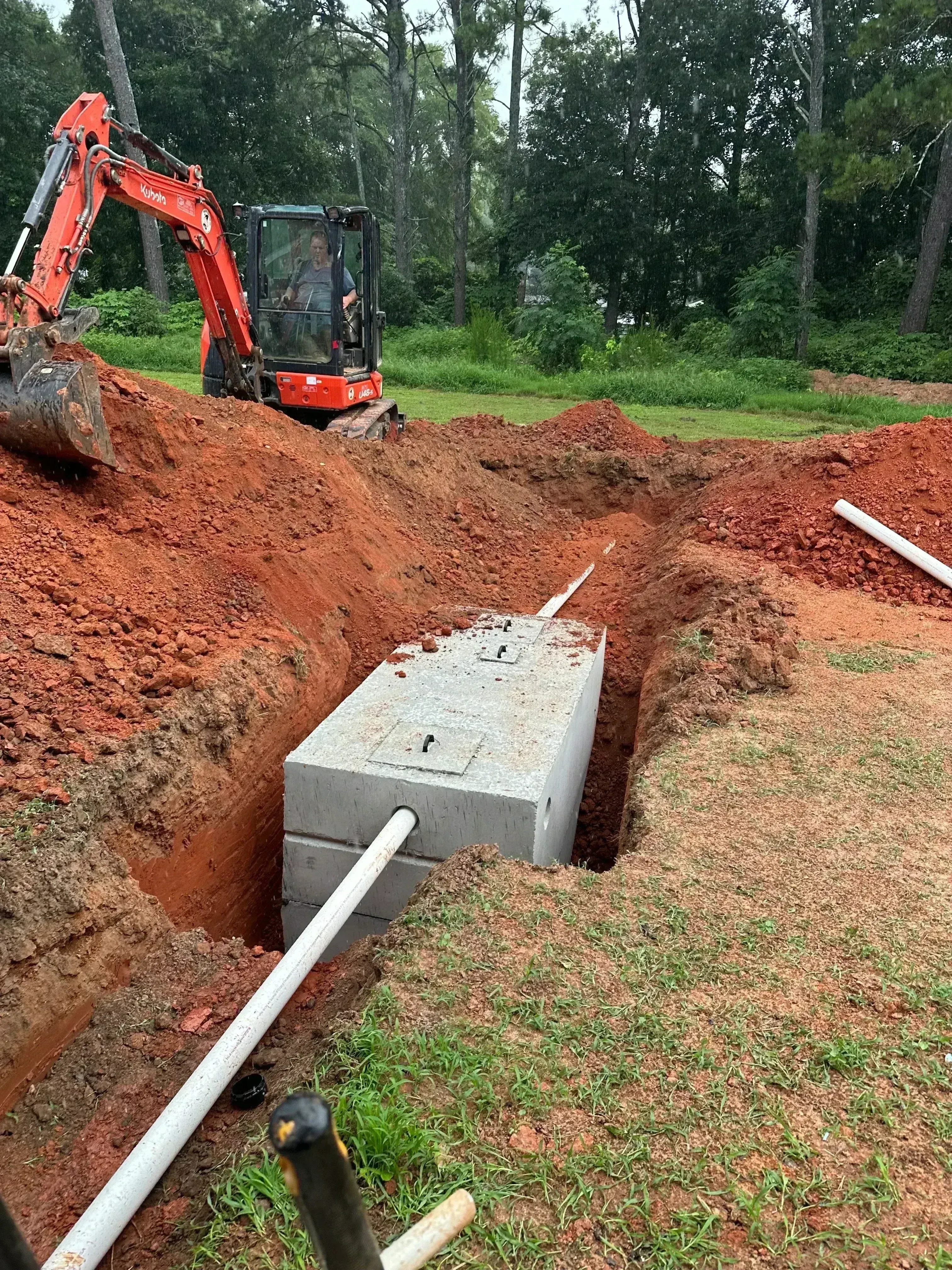Concrete septic tank installed in excavated trench with white PVC pipes and red clay soil