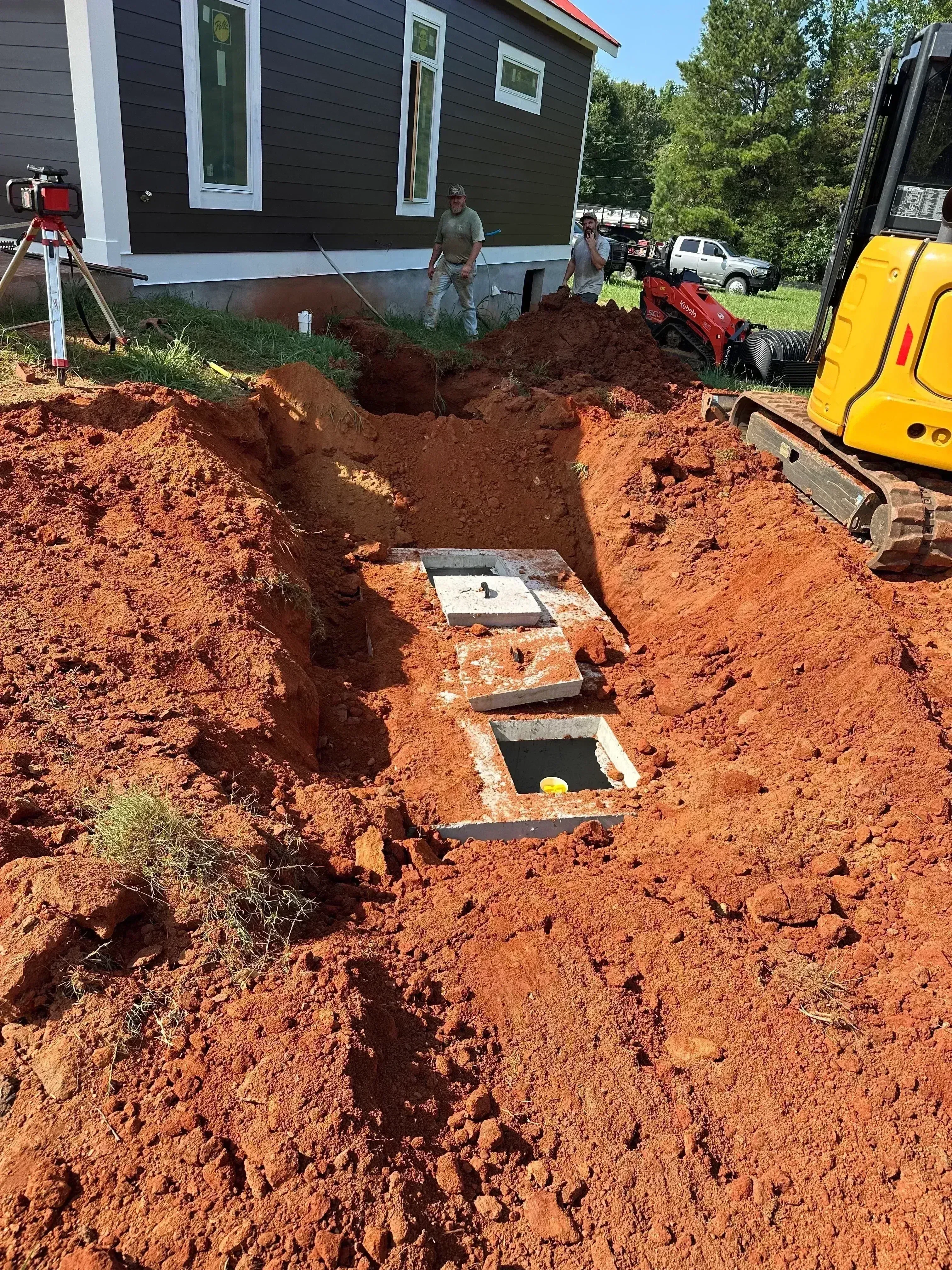 Concrete septic tank installed in excavated pit with clay soil, yellow excavator and red truck on site